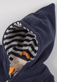 Marineblaues Fleece-Hoodie mit einer spitzen Kapuze, ausgestattet mit einem gestreiften Innenfutter in Weiß und Marineblau, orangenen Akzenten und einem Etikett innen.
