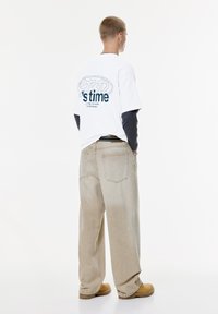 Ung mand står med ryggen til, iført beige brede bukser, brune støvler, lagdelte sorte og hvide graphic T-shirts, hænderne i lommerne.