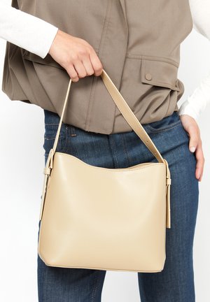 Sac à main en cuir beige avec une texture lisse, un haut arrondi et une sangle ajustable, tenu par une main. Porté avec un jean bleu foncé et un blazer clair.