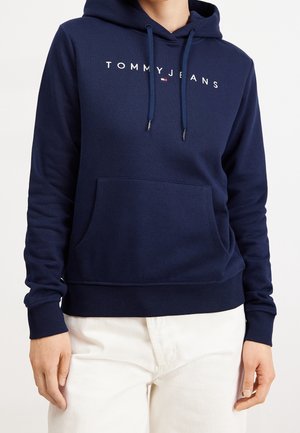 Osoba ubrana w granatową bluzę z kapturem Tommy Jeans z przednią kieszenią i białe spodnie, stojąca na jednolitym tle.