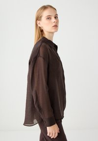 Blusa marrone trasparente con colletto a punta, maniche ampie e design floreale ricamato sul davanti; abbinata a pantaloni coordinati.