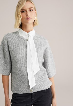 Sivý, rebrovaný pletený sveter s krátkymi rukávmi, ktorý má biely golier so zaväzovaním a zapínanie na gombík v dolnej časti. Viditeľné čierne džínsy.