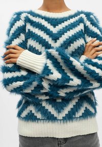 Puszysty sweter w kolorze szmaragdowym i białym z wzorem w zygzaki, przeszyty ściągaczami i o fakturowanym wykończeniu. Posiada okrągły dekolt i miękką tkaninę.