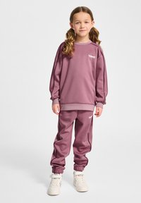 Paarse sweatshirt en bijpassende joggers gemaakt van zacht materiaal, met elastische boorden en een klein logo op de borst en dij. Witte sneakers.