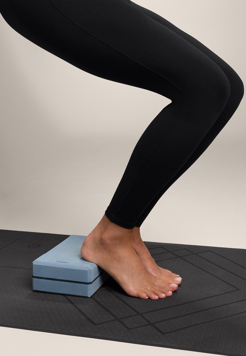 Vaaleansininen vaahtomuovinen joogalohko, suorakulmainen muoto, sijoitettuna jalan alle. Alla musta joogamatto, jossa on kuvioitu pinta.