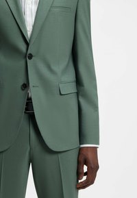 Homme portant un costume ajusté vert clair avec des boutons noirs, associé à une chemise blanche et une ceinture noire, main détendue sur le côté.