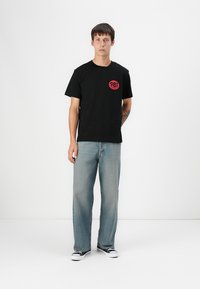 Tricou negru din bumbac cu un grafic circular proeminent roșu având numărul "981". Asociat cu jeans denim deschiși la culoare și adidași negri.