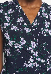 Blommig omslagstopp i marinblått tyg med vita och lila blommor, med en plisserad design och en V-ringning för extra detalj.