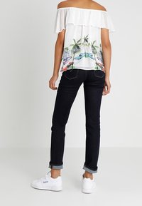 Haut blanc à épaules nues avec des manches volantées, arborant un imprimé coloré de palmiers et de voitures ; associé à un jean foncé et des baskets blanches.