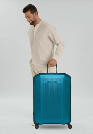 Hombre con atuendo beige sosteniendo el asa de una gran maleta azul SMARTBOX con cuatro ruedas, de pie frente a un fondo claro y sencillo.