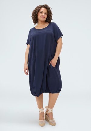 Frau mit lockigem Haar, die ein locker sitzendes dunkelblaues Kurzarmkleid und beige Keilsandalen mit Knöchelriemen trägt, steht vor einem schlichten Hintergrund.
