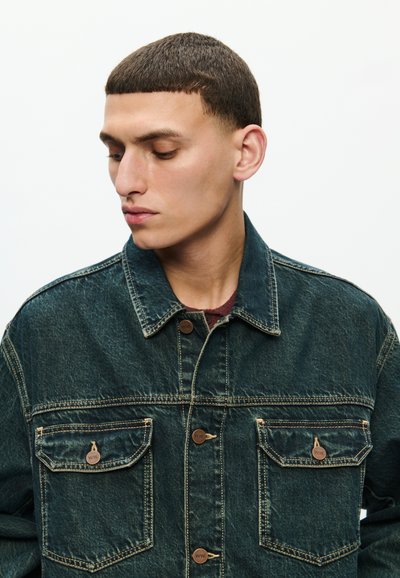 Giovane uomo con capelli corti e scuri che guarda verso sinistra con la testa leggermente abbassata, indossa una giacca di denim scuro con tasche sul petto chiuse da bottoni, su uno sfondo chiaro e uniforme.