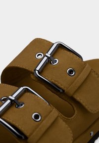 Καφέ σανδάλια από σουέτ με δύο φαρδιές λουρίδες, διακοσμημένα με ασημένιες αγκράφες και στρογγυλές μεταλλικές τρύπες, που αναδεικνύουν έναν μινιμαλιστικό σχεδιασμό.