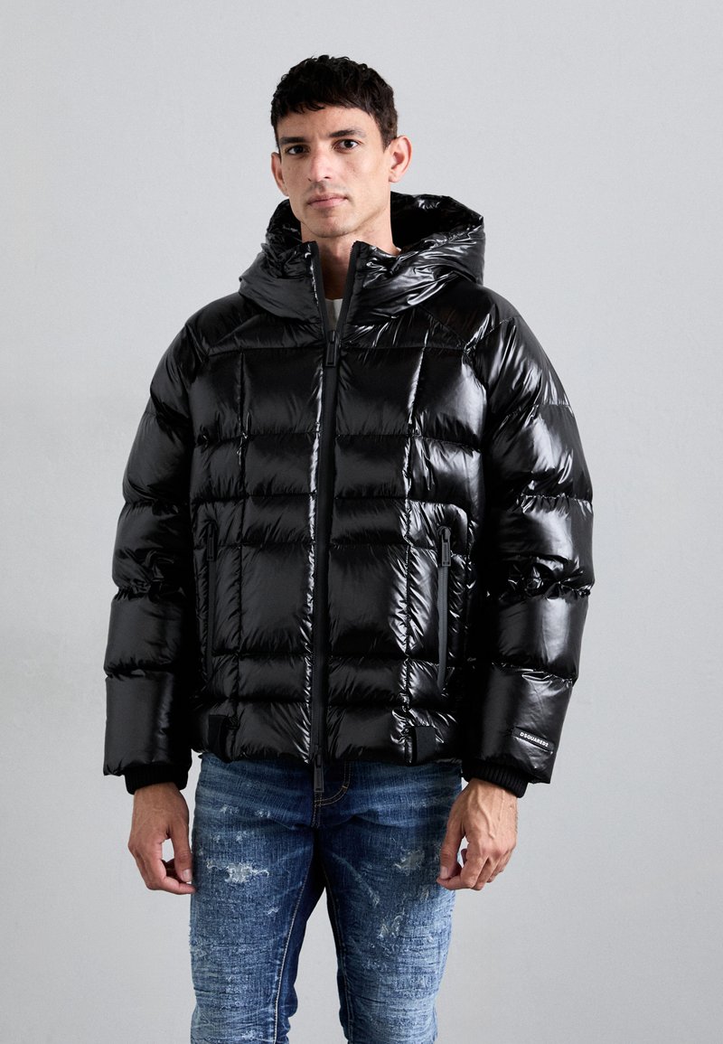 Giubbotto imbottito nero con finitura lucida, dotato di cappuccio, chiusura con zip e design trapuntato. Indossato sopra jeans blu strappati.