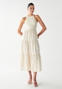 Nainen seisoo kädet selän takana, päällään beige säärystinmidimekko ilman hihoja ja valkoiset avokkaat tasaisella valkoisella taustalla.