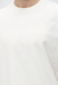 Κοντινό ενός ατόμου που φοράει ένα απλό λευκό t-shirt με λαιμόκοψη και διακριτικό ανάγλυφο κείμενο στο στήθος.