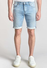 Pantalones cortos de mezclilla azul claro con dobladillos enrollados, que cuentan con un diseño clásico de cinco bolsillos y cierre de botón metálico. Llevados con zapatillas deportivas blancas.