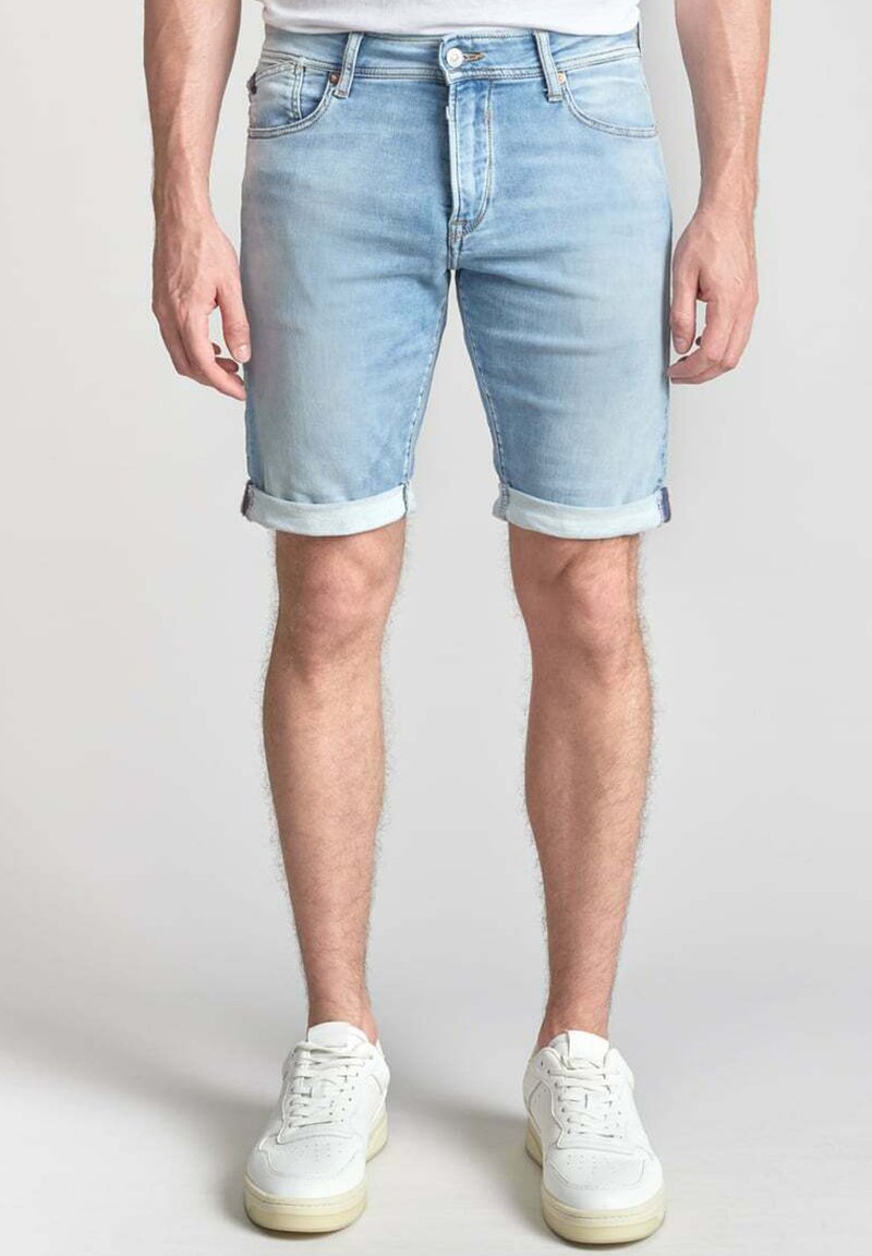 Pantalones cortos de mezclilla azul claro con dobladillos enrollados, que cuentan con un diseño clásico de cinco bolsillos y cierre de botón metálico. Llevados con zapatillas deportivas blancas.