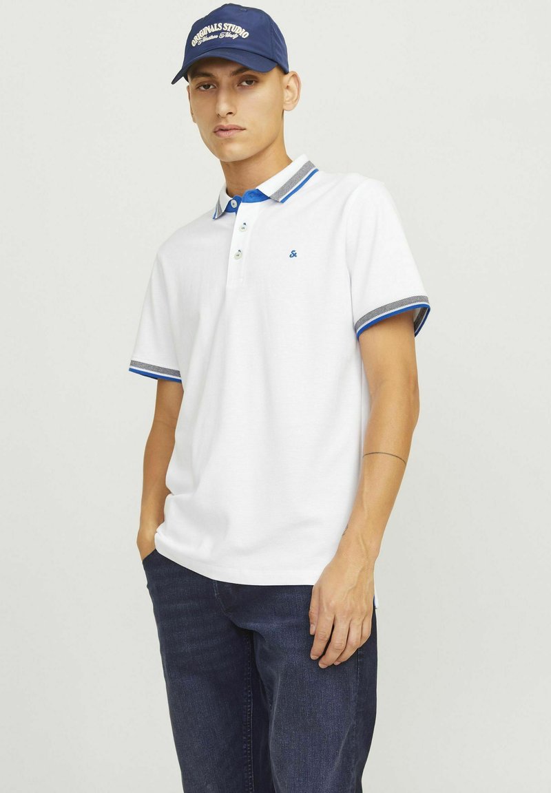 Weißes Polohemd mit blauen und grauen Akzenten am Kragen und an den Ärmeln. Drei-Knopf-Leiste. Kombiniert mit dunklen Jeans.