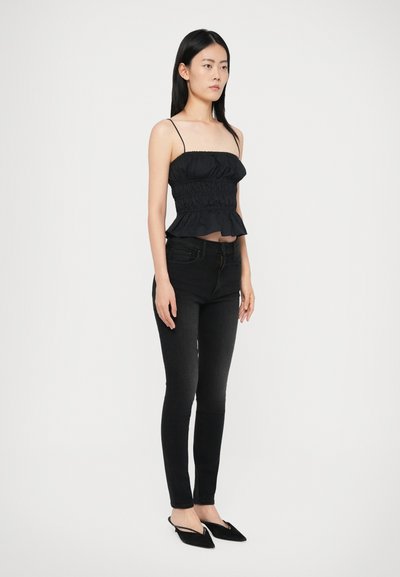 Mulher vestindo um top preto com plissados sem mangas, jeans skinny pretos e sapatos slingback pretos com ponta, de pé contra um fundo branco liso.