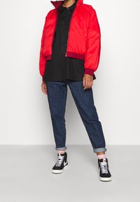 Červená prošívaná bomber bunda s žebrovanými manžetami a lemem, nošená přes černou košili na knoflíky, doplněná o ohrnuté modré džíny a černé tenisky.