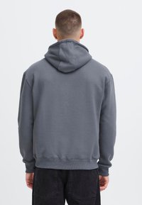 Sweat à capuche gris avec une texture douce, des poignets et un ourlet côtelés, et un détail de couture au dos, porté avec un pantalon foncé.