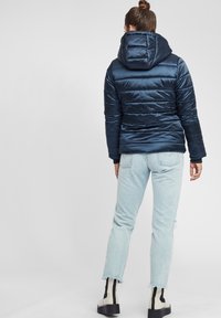 Chaqueta acolchada azul marino con capucha, con un diseño de acolchado horizontal y textura suave. Combinada con jeans desgastados de color azul claro y botas en blanco y negro.