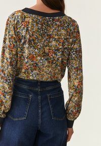 Floral blouse with long sleeves in yellow, red, and blue tones. Features a navy collar and a smooth fabric. Paired with high-waisted dark denim jeans.