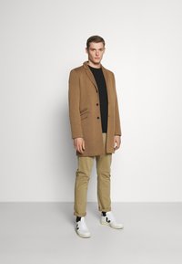 Brauner Mantel mit Reverskragen, einreihigem Design und zwei vorderen Taschen, kombiniert mit einem schwarzen Hemd, beigen Hosen und weißen Turnschuhen.