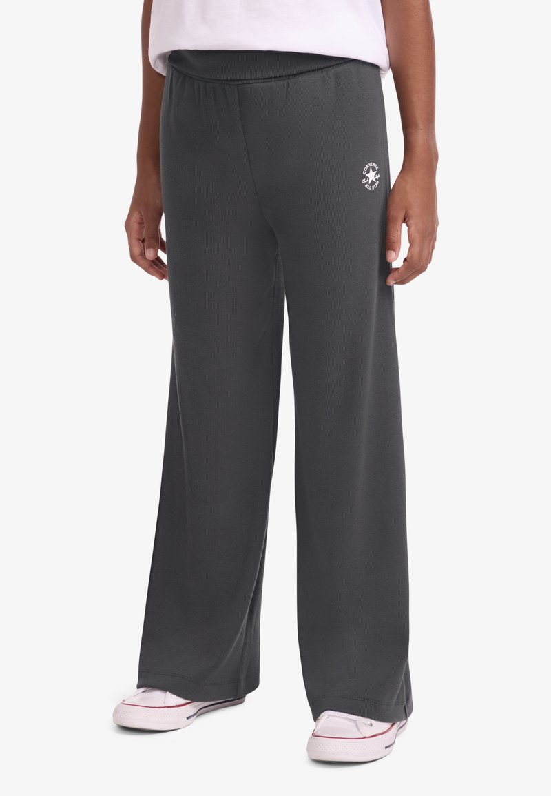 Γκρι φαρδιά φόρμα με απαλό ύφασμα, ελαστική μέση και μικρή λογότυπο στο αριστερό μηρό. Φορώντας με λευκά παπούτσια.