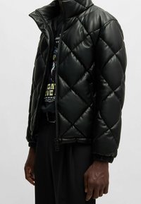 La giacca a vento nera trapuntata presenta un design lucido e imbottito, colletto alto e polsini in elastico. La chiusura è a zip e sono visibili le tasche laterali.