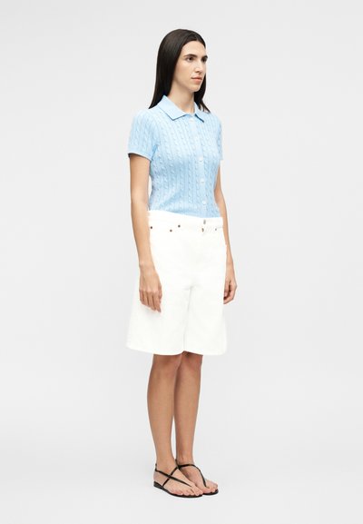 Nő világoskék, rövid ujjú, galléros kötött felsőben, fehér térdig érő rövidnadrágban és fekete pántos lapos szandálban áll.