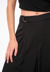 Zwarte geplooide broek met een zilveren ronde hardware detail bij de taille, gemaakt van gladde stof, met een ruime pasvorm en zijslits.