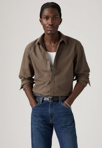 Man met gevlochten haar, gekleed in een bruin overhemd over een witte tanktop, blauwe jeans, een zwarte riem en zilveren sieraden, met zijn handen in de zakken.