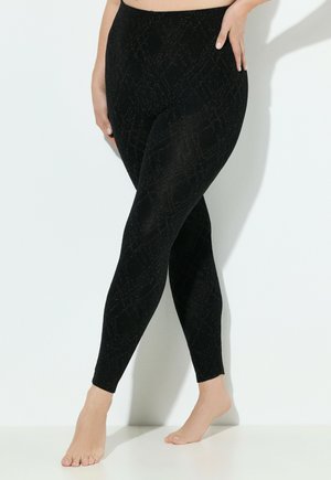 Kvinde-ben iført sorte mønstrede leggings, den ene hånd hvilende på hoften, står barfodet mod en hvid baggrund.