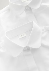 Białe koszule z kołnierzykiem, ułożone w stos, wykonane z gładkiego materiału, z guzikami i metką z napisem "NEXT". Kołnierzyki są zaokrąglone i starannie wyprasowane.