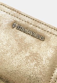 Goldfarbener Geldbeutel mit einer strukturierten, metallischen Oberfläche. Mit dem silbernen "Stradivarius"-Logo. Genähte Kanten und glatte Konturen.