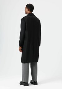 Lange zwarte jas met een rechte snit, voorzien van een kraag en een enkele split aan de achterkant. Gecombineerd met grijze broek en zwarte schoenen.