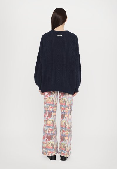 Pull en maille oversize bleu marine avec un motif de câbles texturé ; associé à un pantalon large à motif graphique multicolore et des bottes à talons noires chunky.