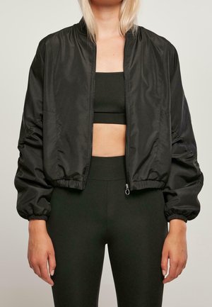 Vrouw draagt een zwart cropped bomberjack met rits over een zwarte sportbeha en een high-waisted legging tegen een effen achtergrond.