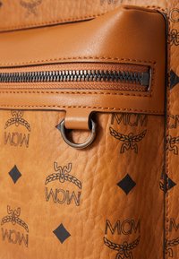 Détail d'un sac en cuir brun avec une fermeture éclair noire, un anneau en métal et un motif logo en relief avec des accents en forme de diamant sur la surface.