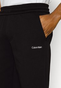 Sorte joggebukser med en glatt tekstur, med en Calvin Klein-logo i hvitt på hoften og en sidelomme. Elastisk livlinje.