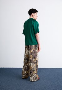 Πράσινο κοντομάνικο πουκάμισο με υφή πλέγματος, συνδυασμένο με καφέ παντελόνια φαρδιάς γραμμής με καμουφλάζ και τσέπες. Ορατά κόκκινα υποδήματα.