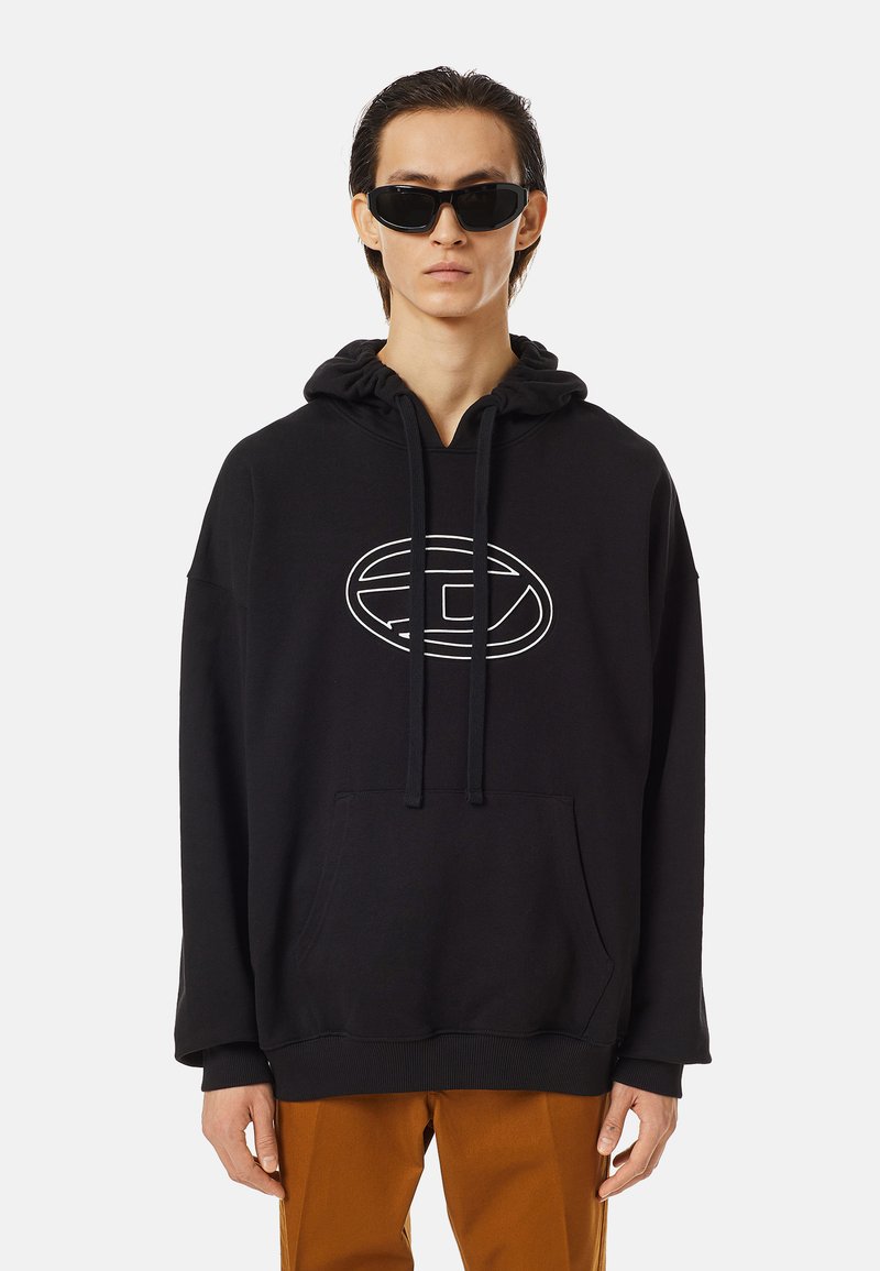 Jeune personne portant un sweat à capuche noir avec un logo blanc, un pantalon marron et des lunettes de soleil noires, se tenant devant un fond blanc uni.