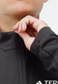 Person's hand pulling up the zipper of a black jacket with visible white text and logo near the bottom right.