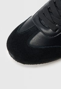 Sapatilha desportiva preta com biqueira em camurça texturada e laterais em couro liso. Apresenta atacadores pretos e costuras, com uma silhueta arredondada.