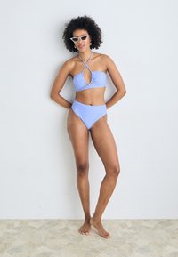 Lyseblå bikini med rynkede detaljer og kryssryggdesign. Høyt liv på bunnene. Modellen står på et strukturert steingulv.