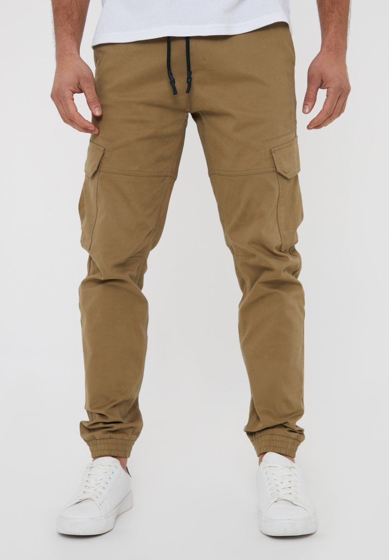 Pantalons cargo en khaki fabriqués en coton résistant, avec des chevilles fuselées, des poignets élastiques et de multiples poches latérales. Association avec des baskets blanches.
