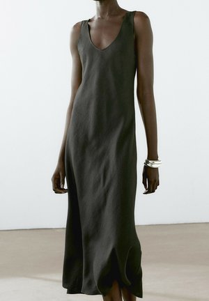 Femme portant une robe sans manches, foncée, longue jusqu'aux chevilles avec un décolleté en V profond, accessoirisée avec des bracelets argentés au poignet droit.