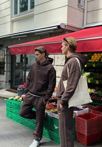 To modeller har matchende mørkebrune hettegensere og joggebukser med en logo. En lysfarget totebag holdes av den ene. Bakgrunnen viser ferske råvarer.